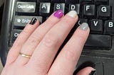 Close up photo of a hand resting on a keyboard. The fingernails are painted in the colours of the asexual flag: black, grey, white, and purple.