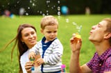 Two young parents play with their toddler with one blowing bubbles and the other holding the child.
