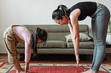 A mother and daughter stretch together, standing and touching their toes, in front of a sofa.