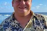 Image of a Keith, a white mane, smiling. He is in Hawaii so you can see the ocean and waves behind him.