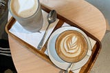 A latte and a cappucino with spoons on the side on a tray on top of a small side table on concrete floor.