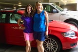 Isabel is 13 and wearning a purple T-shirt and pink shorts. I’m beside her in a blue sleevelss top and navy shorts. We stand in fron of our red Volvo wagon, Ruby, in a parking deck. We are smiling.
