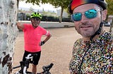 Two men stand in cycling helmets and sunglasses beside their bikes in a village square in France