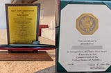 Two recognition plaques from the U.S. Government. The one on the left is from 1965 and is made from wood and metal. The one on the right is from 2001 and is a printed sheet of paper in a plastic folder.