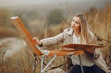 Young woman leaning over a painting box stand, painting a picture of nature outside in a grassy field