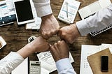 Team members are fist-bumping over a desk of paper and supplies to show they work together.