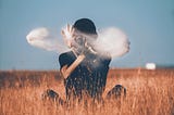 Photo of a man sitting on brown grass and playing with smoke. The man’s face is covered with smoke