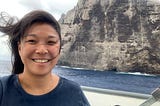 Jiny Kim, wearing a navy blue shirt, stands on a boat with Nihoa in the background.