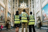 Three people stand, wearing high vis vests with a Night Club logo, with their backs to us in a grand hallway.