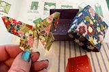 A hand holding a colorful origami butterfly with origami boxes to the right of the hand.