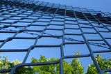 A chain link prison fence with the sky and bushes visable on the other side of it.