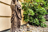 A stand stone statue of Guanyin next to a philodendron. A nine-stone cairn sits at her feet in honor of the author’s mother on her 108th birthday.