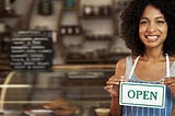 a black woman with an “open” sign on her chest, in front of a coffee shop