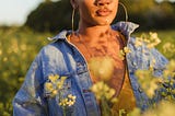 A young woman stands in a field of yellow flowers and stares off into the distance.