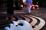 In the front of the photo, a white mask sits on a bronze street bench. The mask depicts a sad face with a single black teardrop. The background is a cityscape at dusk. A figure in white is walking away from the mask.