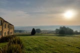 Sun rising over County Durham. A rough lawn of grass and reeds, with trees and rolling hills in the background. A stone hotel building, 2 stories high, is down the left side of the picture.