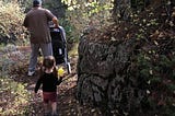 Grandpa pushes a stroller on a hike in the woods with a toddler trailing behind.