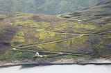 an image of a hill with a zigzag path carved into it, meeting a lake and a small building at the bottom.