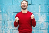 Man in red shirt cheers jubilantly in front of a blue brick wall