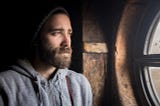 A bearded man wearing a hat looks out the window with a sad expression on his face