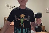 Author smiling while holding a dumbbell in his left hand, wearing a black t-shirt with the words ‘GYM RAT’ and a cartoon design of a rat lifting weights. He is standing in a bedroom with a wooden bed frame and decorative wall art in the background.