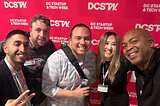 A group of five people posing and smiling at DC Startup & Tech Week in front of a red backdrop with the event logo. From left to right, a man in a suit makes a peace sign, the second person (wearing a Brady UX name tag) stands in a graphic tee, the third person is holding a drink and smiling, the fourth person is a woman with long hair, and the fifth person is smiling broadly. They appear to be enjoying the networking atmosphere of the event.