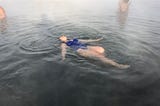Woman on her back in a blue bathing suit with leg outstretched and arms at her sides in the middle of a hot spring.