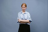 A young woman with red hair wearing a blue, button-up shirt and black skirt with a determined expression.