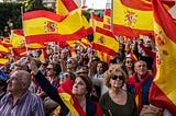 Photo of people waving Spanish flags during a protest demanding general elections and against Pedro Sanchez’s Government after motion of censure, in Madrid, Spain. The crowd is looking towards the left, with a few people looking towards the right in the bottom corner of the photo.