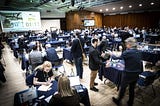 A large crowded room filled with tables for various meetings, and a projector reflects a clock ticking to direct the attendees when to move to their next meeting.