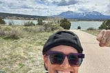 CC dressed in a black long sleeve shirt and black cap with sunglasses smiling and doing a strong arm selfiie in the camera with a background of mountains and a lake.