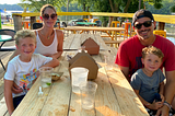 Zach sitting at a picnic table with his partner and two children, smiling in the sun.