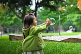 Asian toddler in a green oat playing with a bubble in a park, Jessie Waddell, Medium