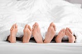 Three pairs of feet sticking out from under the bed sheets