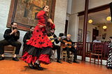 A female flamenco dancer in a frilly red and black dress dances in front of four male singers and guitarists.