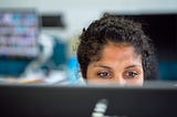 A woman with curly black hair that is pulled back, sits in a computer lab with her eyes peeking over a computer screen.