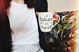 Dark haired woman in a white shirt and black jacker holding a coffee mug with the slogan “Believe in Yourself”