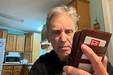 An older man standing in a kitchen, looking intently and somewhat frustrated or even angry at his smartphone, which is in a leather wallet-style case. The background shows wooden cabinets, a microwave, and a bright overhead light.