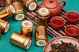 Wuyi rock tea, a specialty of ITEAMO, is displayed in brass metal cannisters, beside the dried roasted leaves, and several cups of the brew served in a bright red tea set.