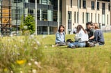 A group of four students sit cross legged on grass outside the Michael Marks building. They are all dressed in light summer clothing. and are smiling and in conversation. There is greenery in the foreground out of focus. The building is modern architectural style.