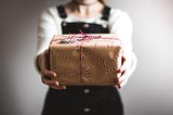 An anonymous woman holds out a package gift-wrapped in brown paper with a candy cane design and a real candy cane tied to the top with red and white string.