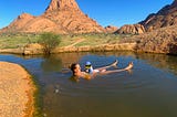 A woman floats in a pond with her child on top. Green grass is in the background.