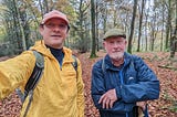 Two men (my dad and me) wearing waterproof coats and hats in a wood along the route of the South Downs Way. It is autumn and the ground is covered with red and brown leaves.
