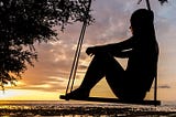 The silhouette of a woman sitting alone on an outdoor swing in the dark with the sunset behind her.