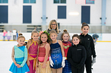 Martine De La Torre poses with a group of young skaters on the ice. Some are wearing colorful skating costumes and others are wearing black jackets and leggings. De La Torre stands behind the skaters smiling. She is a white woman with shoulder-length blonde hair and glasses.