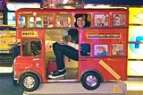 The author (a white, dark-haired femme person) in a minute red, double-decker bus meant for children to sit in. It is side-on and she is waving at the camera.