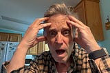 An older man indoors, wearing a plaid shirt and a smartwatch, is holding his head with both hands and looking directly at the camera with a shocked or distressed expression.