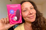 Woman smiling while looking into the camera while holding up a pink box containing a vibrator next to her face.