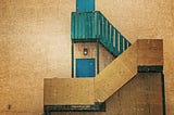 Side of an old building in brownish particle board with a wooden staircase leading to a green staircase and a blue door.