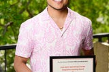 Young, caramelicious man holding an award frame wearing pink and white in front of greenery.